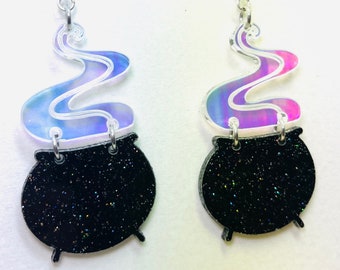 May include: A pair of iridescent black cauldron earrings with a purple and blue iridescent smoke detail. The earrings are made of acrylic and have silver hooks.