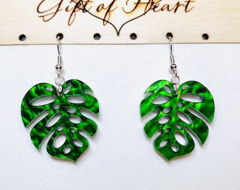 May include: Green monstera leaf earrings with a marbled pattern and silver hooks. The earrings are hanging from a wooden surface with the text "Gift of Heart" above them.