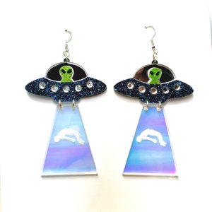 May include: A pair of dangle earrings featuring a blue and black glitter UFO with a green alien inside. The bottom of the earrings are iridescent blue and feature a white silhouette of a person falling.