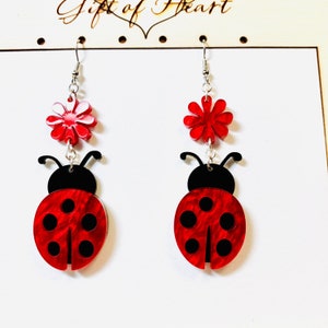 May include: A pair of red ladybug earrings with black spots and a red flower charm. The earrings are made of acrylic and have silver hooks.