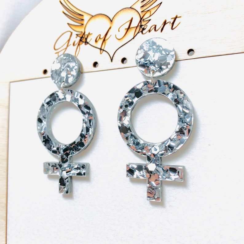 May include: Silver glitter earrings shaped like the female gender symbol. The earrings are on a wooden display card with the text "Gift of Heart".