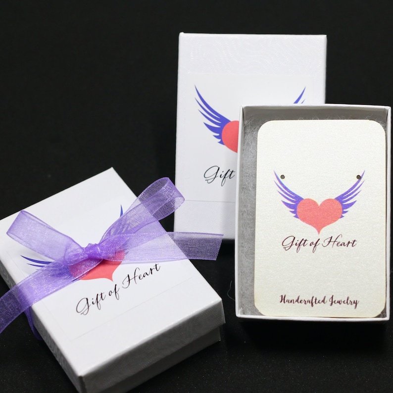K&ouml;nnte beinhalten: Zwei wei&szlig;e Geschenkboxen mit lila Band und einem pinkfarbenen Herz mit Fl&uuml;geln-Logo. Das Logo lautet "Gift of Heart Handcrafted Jewelry".