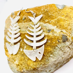 May include: A pair of white fish bone earrings with silver hooks. The earrings are made of a white, marbled material and are shaped like fish skeletons.