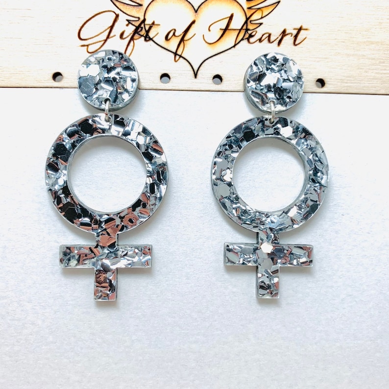 May include: A pair of silver earrings shaped like the female symbol. The earrings are made of a glittery material and have a textured finish.