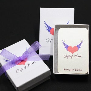 May include: Two white gift boxes with purple ribbon and a pink heart with blue wings logo. The text on the boxes reads "Gift of Heart" and "Handcrafted Jewelry".