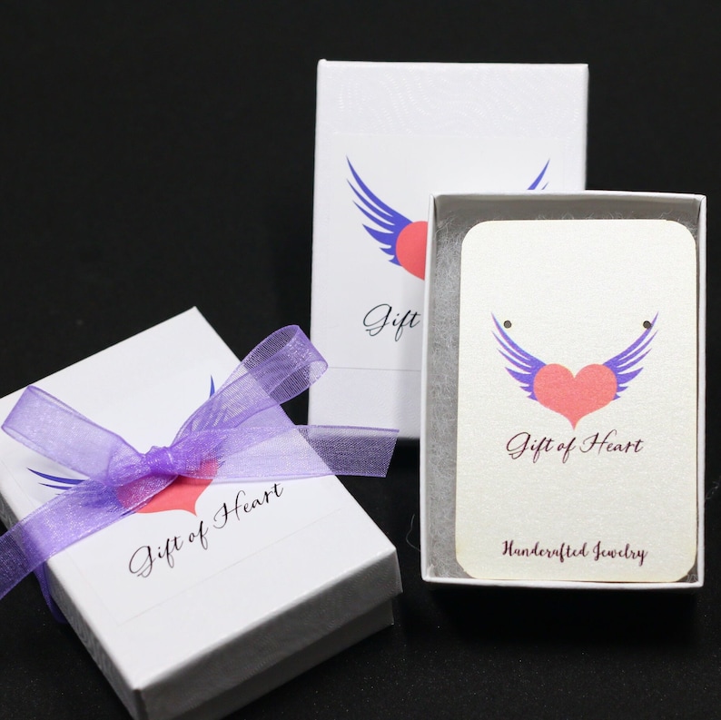 May include: Two white gift boxes with purple ribbon and a pink heart with blue wings logo. The text on the boxes reads "Gift of Heart" and "Handcrafted Jewelry".
