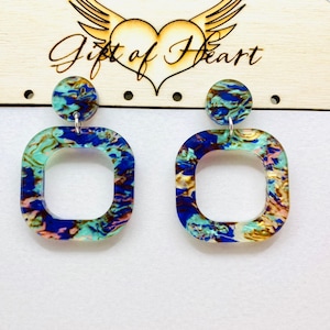 May include: A pair of square-shaped earrings with a blue, green, gold, and pink marbled design. The earrings have a smaller round disc attached to the top.