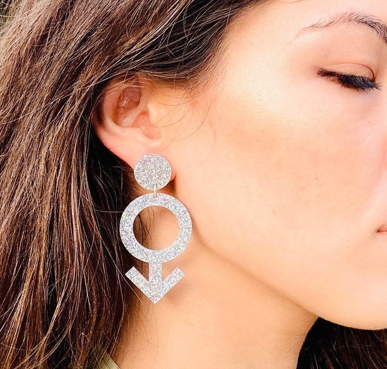 May include: Silver glitter earrings shaped like a male symbol with a circle and arrow. The earrings are dangling and have a small circle at the top.
