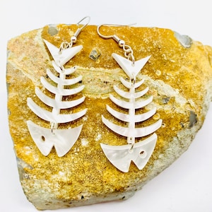 May include: A pair of white fishbone earrings with silver hooks. The earrings are made of a pearlescent material and have a delicate, whimsical design.