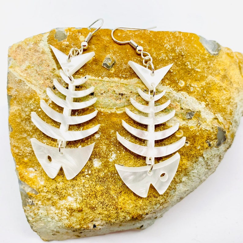 May include: A pair of white fishbone earrings with silver hooks. The earrings are made of a pearlescent material and have a delicate, whimsical design.