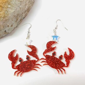 May include: A pair of red glitter crab earrings with a silver star charm. The earrings are on a white background.