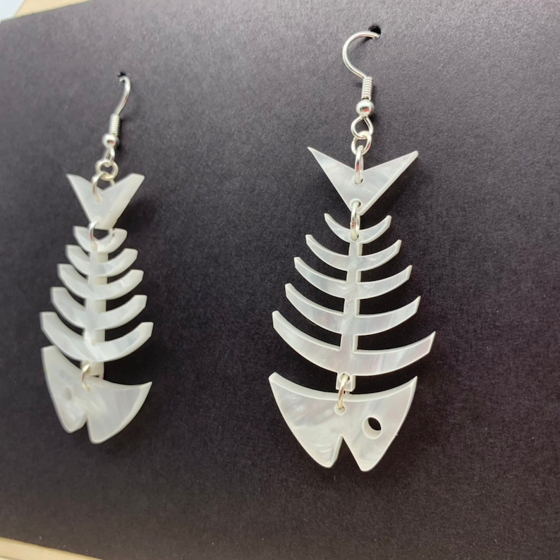 May include: A pair of white fish bone shaped earrings with silver hooks. The earrings are made of a translucent material with a pearlescent finish.