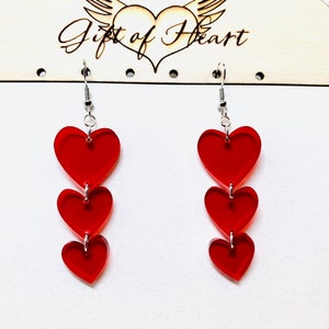 May include: A pair of dangle earrings with three red heart-shaped charms hanging from silver hooks. The earrings are displayed on a white card with the text "Gift of Heart".