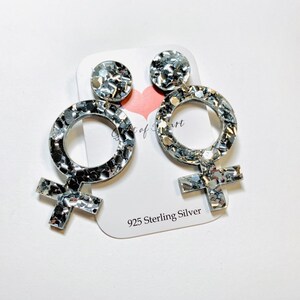 May include: A pair of silver glitter earrings shaped like the female symbol. The earrings are made of 925 sterling silver.