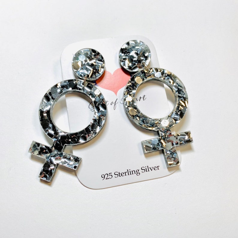 May include: A pair of silver glitter earrings shaped like the female symbol. The earrings are made of 925 sterling silver.