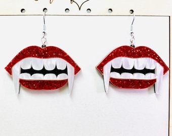May include: A pair of dangle earrings featuring red glitter lips with white pearlescent fangs. The earrings hang from silver-toned metal hooks and are displayed on a white card with the text Gift of Heart printed in black at the top.