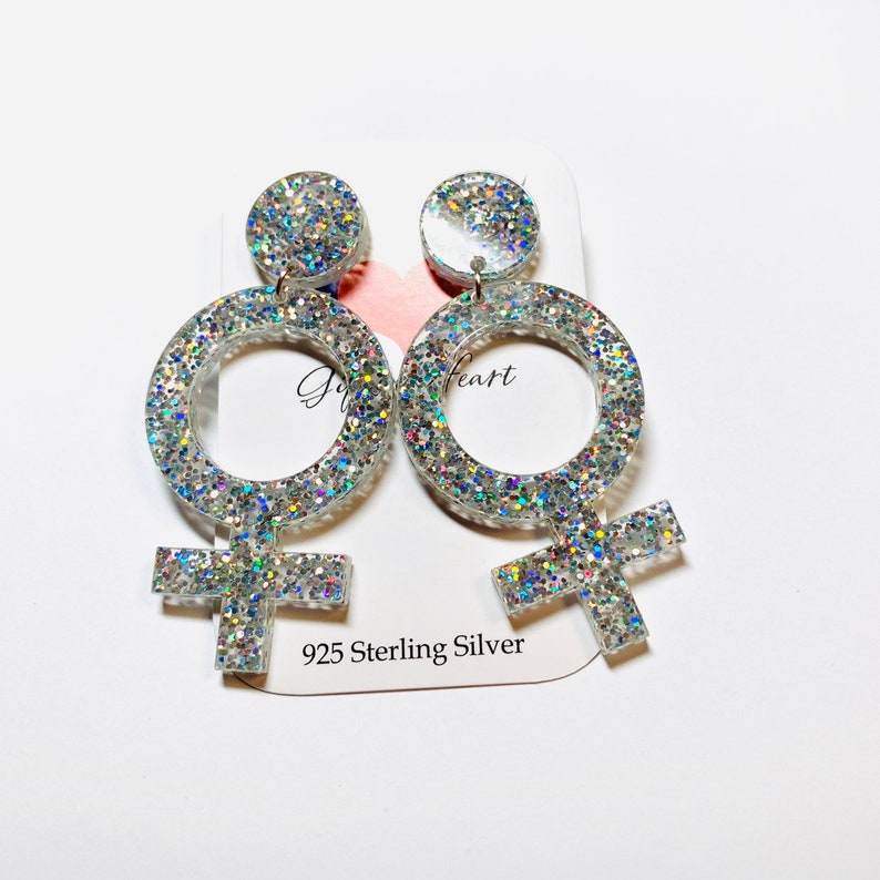 K&ouml;nnte beinhalten: Ein Paar silberne Glitzerohrringe in Form des weiblichen Geschlechtszeichens. Die Ohrringe sind aus 925er Sterlingsilber.
