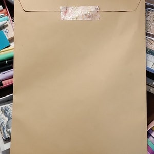 Puede incluir: Un sobre de papel marrón con un sello decorativo de cinta con estampado dorado y blanco. El sobre está sobre una mesa con varias cintas washi de colores y otros materiales de manualidades.