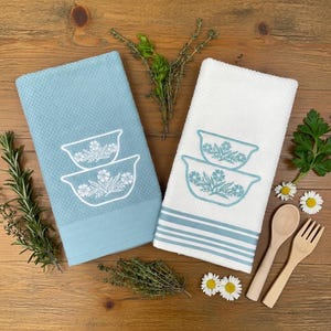 May include: Two kitchen towels, one light blue and one white, with embroidered floral bowl designs. The blue towel has a solid bottom border, while the white towel has blue stripes. Wooden spoon and fork, herbs, and daisies are arranged around the towels.