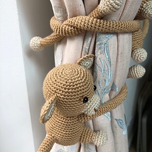 May include: Three crocheted monkey curtain tiebacks in brown and cream colors. The monkeys are holding onto the curtain with their tails wrapped around it.
