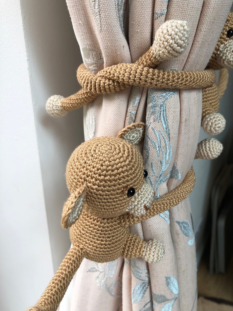 May include: Three crocheted monkey curtain tiebacks in brown and cream colors. The monkeys are holding onto the curtain with their tails wrapped around it.