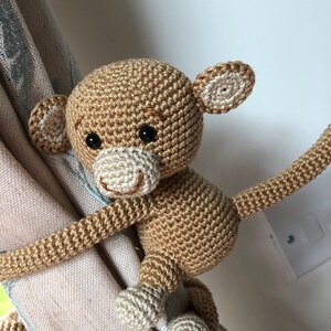 May include: A crocheted monkey toy with brown fur, white ears, and a white tail. The monkey is holding onto a curtain with its tail and one hand.