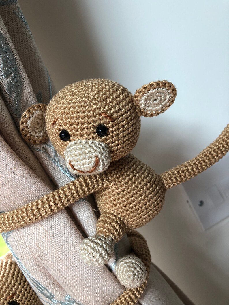 May include: A crocheted monkey toy with brown fur, white ears, and a white tail. The monkey is holding onto a curtain with its tail and one hand.