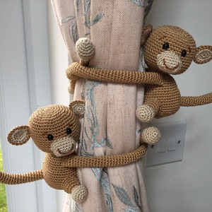 May include: Two brown crocheted monkey curtain tiebacks holding a beige curtain with a floral pattern.