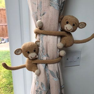 May include: Two crocheted monkey curtain tiebacks in brown, holding a beige curtain with a floral pattern. The monkeys are holding the curtain with their arms and legs.