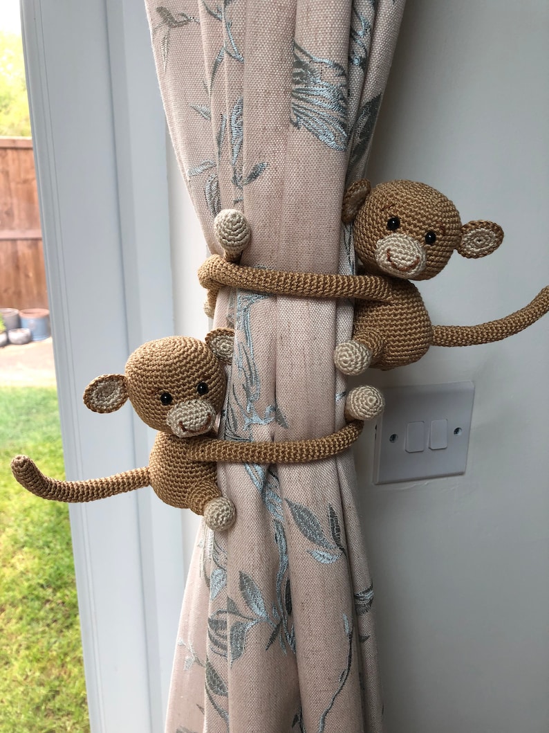 May include: Two crocheted monkey curtain tiebacks in brown, holding a beige curtain with a floral pattern. The monkeys are holding the curtain with their arms and legs.