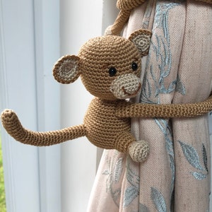 May include: A crocheted monkey toy holding a curtain tieback. The monkey is brown with black eyes and a white mouth. The tieback is made of brown yarn and is wrapped around the curtain.
