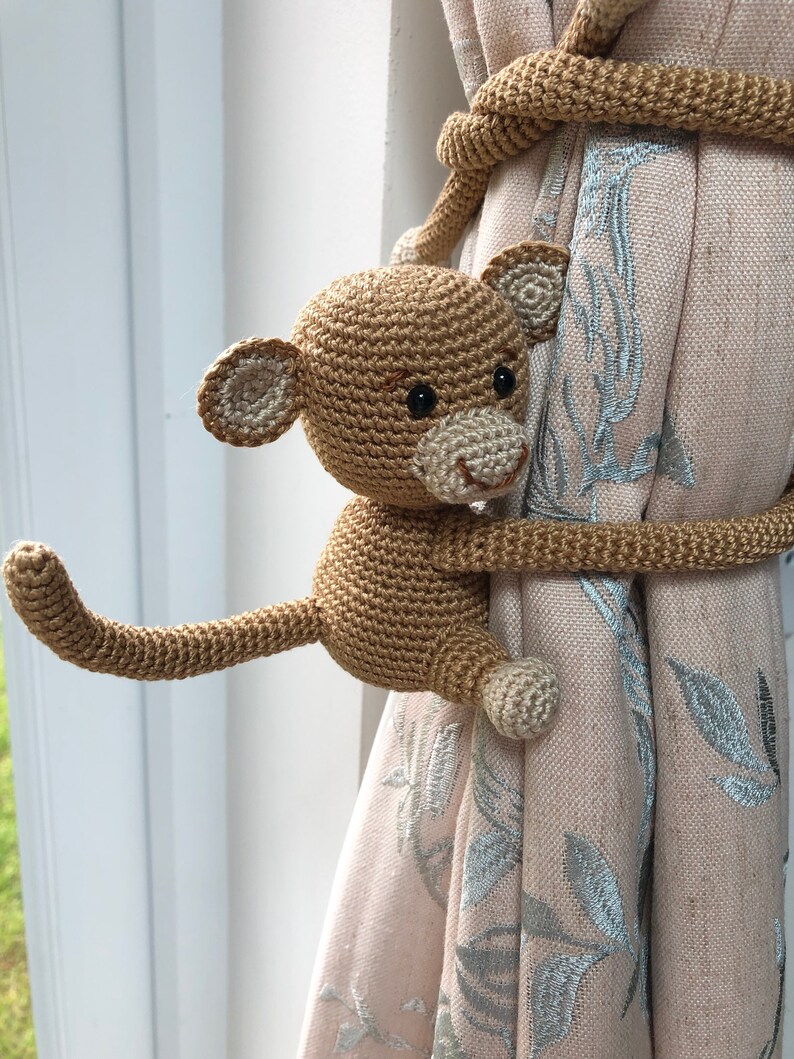May include: A crocheted monkey toy holding a curtain tieback. The monkey is brown with black eyes and a white mouth. The tieback is made of brown yarn and is wrapped around the curtain.