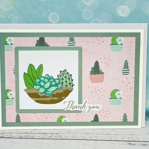May include: A handmade greeting card featuring a watercolor illustration of succulents in a brown pot. The card has a pink background with cactus illustrations and the words "Thank you".