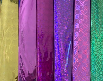 May include: Six rolls of iridescent wrapping paper. The rolls are arranged vertically and are all different colors and patterns. The colors include gold, purple, and green. The patterns include circles, diamonds, and a geometric pattern.