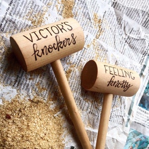 May include: Two wooden mallets with the phrases "VICTOR'S knockers" and "FEELING knotty" written in black script. The mallets are light brown and rest on a newspaper-covered surface with scattered grains.
