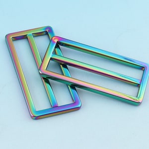 Puede incluir: Dos deslizadores de hebilla de cinturón de metal iridiscente. Los deslizadores son rectangulares con una sola barra en el centro. Los deslizadores son un arcoíris de colores.