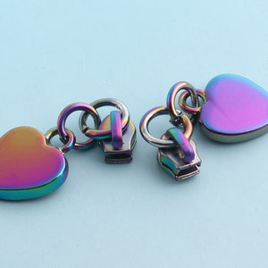 May include: Three iridescent heart-shaped zipper pulls with metal rings. The zipper pulls are a rainbow of colours, including pink, purple, blue, and green.