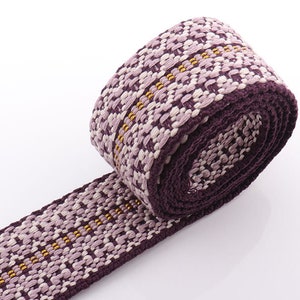 May include: Rolled-up woven fabric with a repeating pattern in shades of purple, lavender, and gold. The fabric has a dark purple border and a textured appearance, suggesting a decorative or craft material.