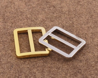 Könnte beinhalten: Zwei Metall-Gürtelschließen, eine goldfarben und eine silberfarben. Beide sind rechteckig mit einem einzigen, mittig angeordneten, horizontalen Balken.