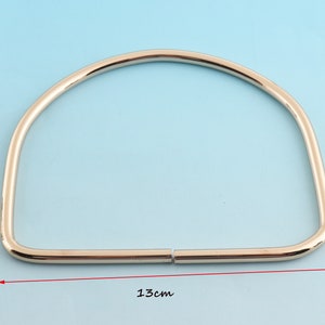 May include: A shiny, gold-colored metal purse handle, semi-circular in shape. The handle measures 5.1 inches across. The handle is made of two pieces that connect in the middle. The background is a light blue color.