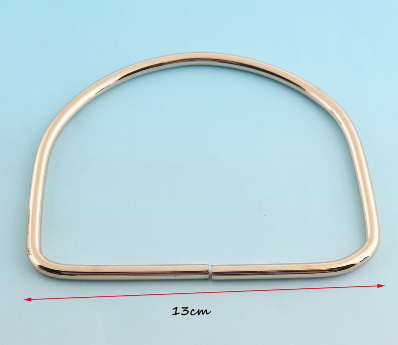 May include: A shiny, gold-colored metal purse handle, semi-circular in shape. The handle measures 5.1 inches across. The handle is made of two pieces that connect in the middle. The background is a light blue color.