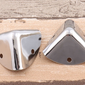 May include: Two silver corner protectors, each with three holes, are shown on a wooden surface.