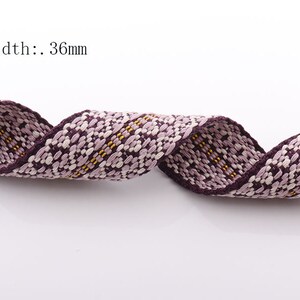 May include: Close-up of a woven ribbon with a floral pattern in shades of purple, white, and yellow. The ribbon is 36mm wide and has a dark purple border. The ribbon is curled and appears to be made of fabric.