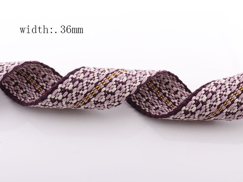 May include: Close-up of a woven ribbon with a floral pattern in shades of purple, white, and yellow. The ribbon is 36mm wide and has a dark purple border. The ribbon is curled and appears to be made of fabric.