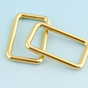 May include: Two gold-colored metal rectangular rings, one slightly larger than the other, are stacked on top of each other.