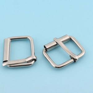Puede incluir: Dos hebillas de cinturón de metal plateado. Una hebilla es de forma cuadrada simple, mientras que la otra tiene un diseño más intrincado con una barra curva.