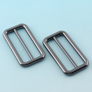 Puede incluir: Dos hebillas deslizantes de metal negro. Las hebillas son rectangulares con un acabado liso y brillante.