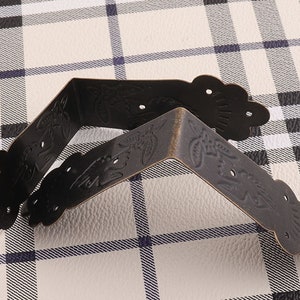 May include: Two black metal corner brackets with an ornate design. The brackets are on a white and black checkered surface.