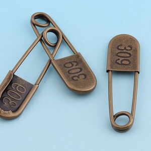 May include: Three antique gold safety pins with the number "303" engraved on each. The pins are on a light blue background.