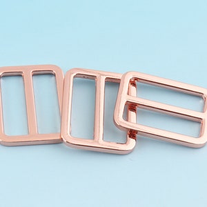 Puede incluir: Tres deslizadores rectangulares de oro rosa con una barra central. Los deslizadores son de metal y tienen un acabado brillante. Están dispuestos sobre un fondo azul claro. Estos deslizadores son adecuados para diversos proyectos de artesanía y costura.